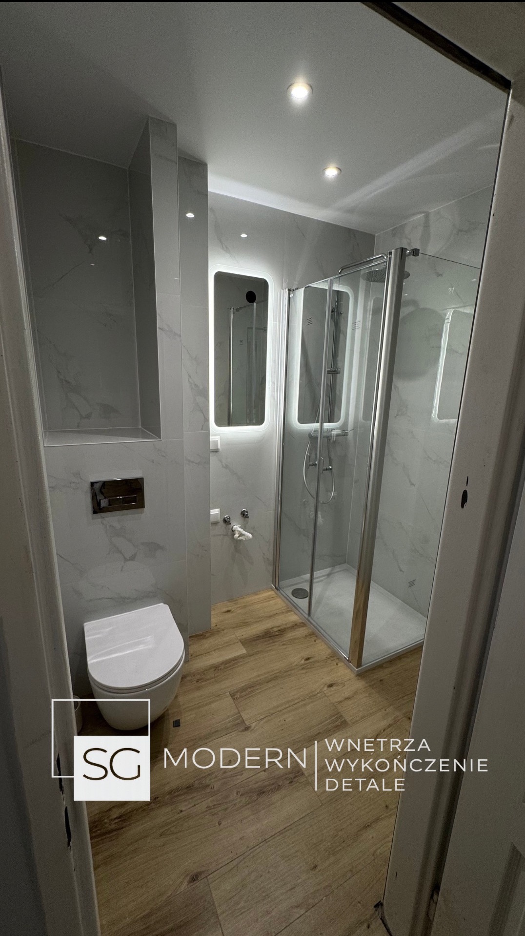Nowoczesna łazienka po remoncie: szare płytki imitujące marmur, drewnopodobna podłoga, kabina prysznicowa i podświetlane lustro. Widoczny fragment logo firmy.