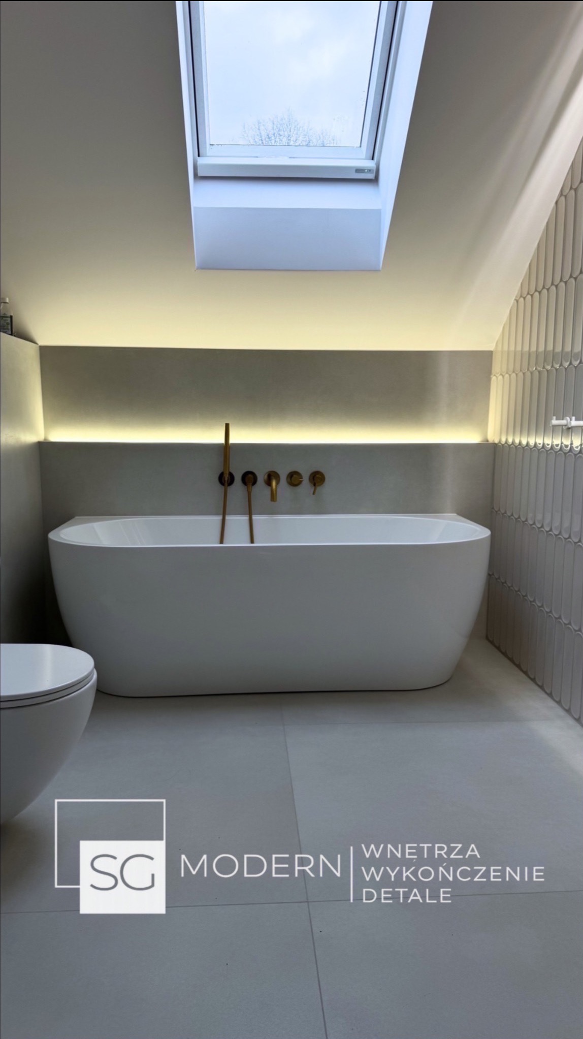 Minimalistyczna łazienka na poddaszu z wolnostojącą wanną, złotą armaturą i oknem dachowym. Ściana wyłożona pionowymi, wąskimi płytkami. Subtelne oświetlenie LED.