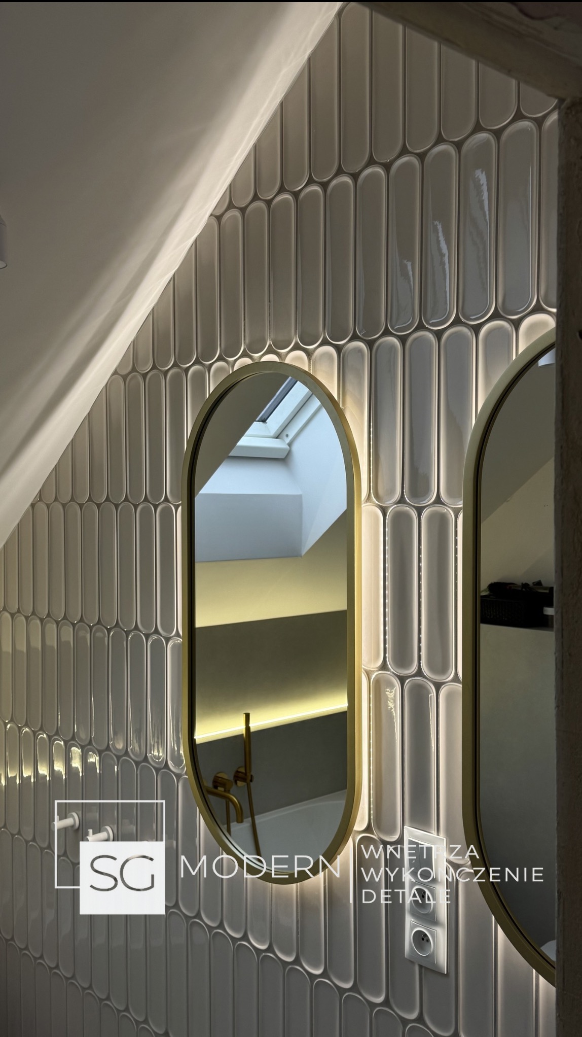 Łazienka na poddaszu z owalnymi, szarymi płytkami, złotymi ramami luster i oświetleniem LED. Widoczne okno dachowe i fragment instalacji. Nowoczesny design.