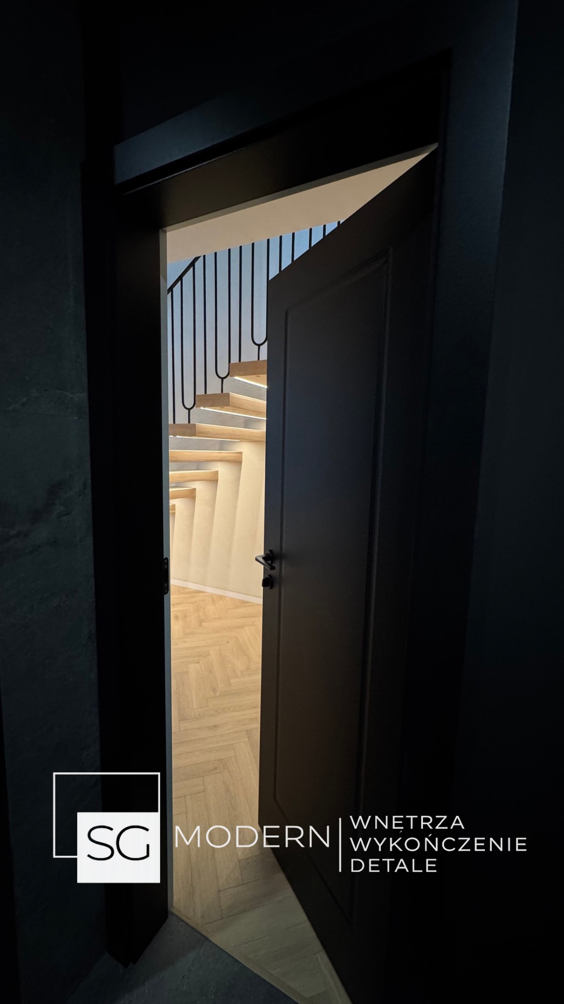 Ciemne drzwi uchylone, ukazujące schody z podświetleniem LED i parkiet w jodełkę. Minimalistyczny design wnętrza, widok z korytarza.