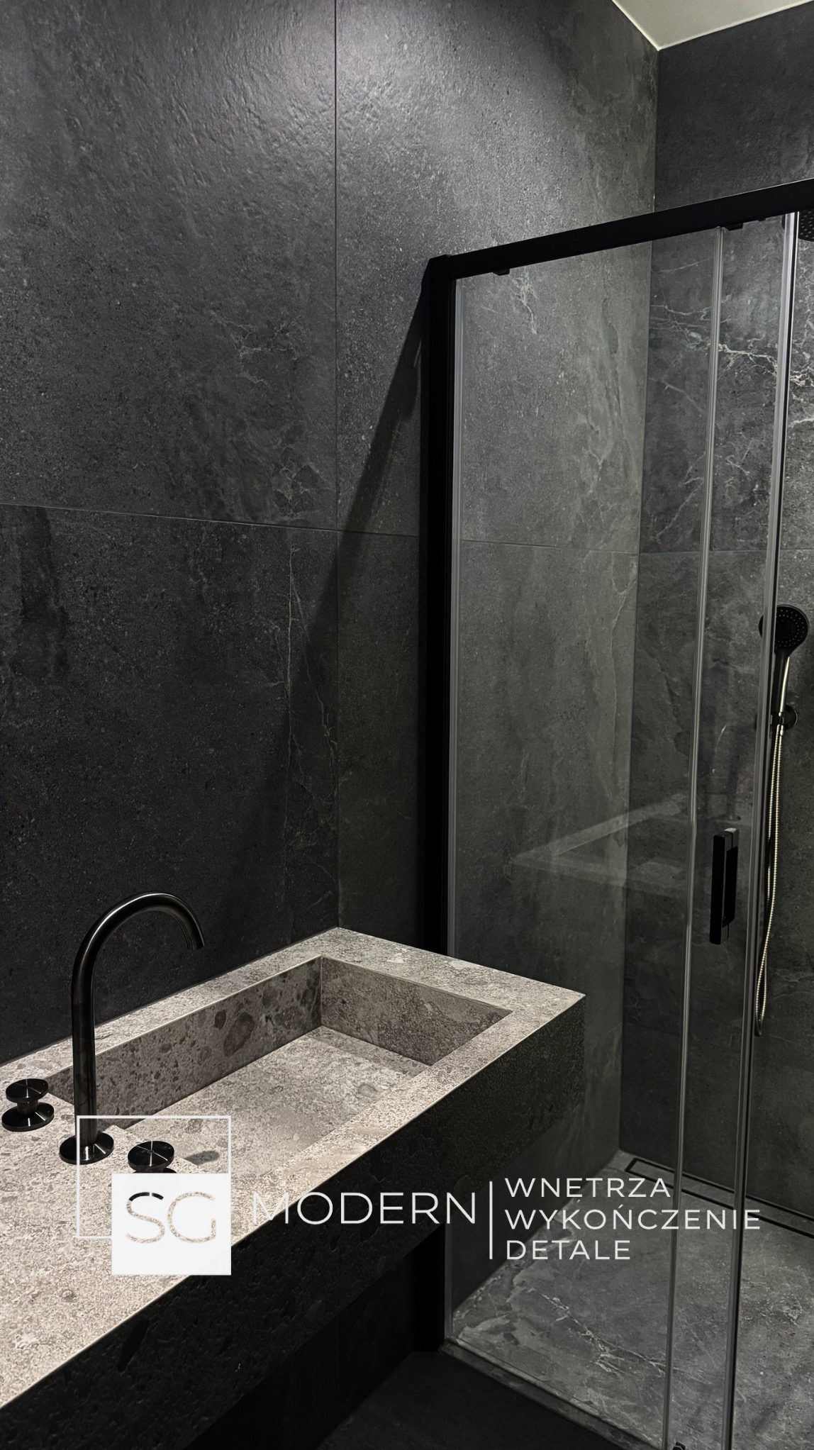 Nowoczesna łazienka z kabiną prysznicową typu walk-in, umywalką z kamienia i czarną armaturą na tle ciemnych, grafitowych płytek. Minimalistyczny design.