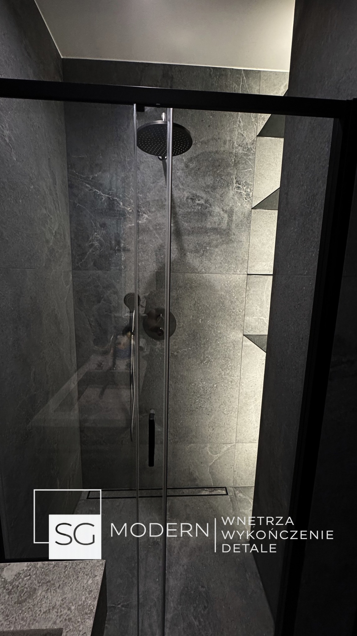 Nowoczesna kabina prysznicowa z czarnymi profilami i deszczownicą, wykończona szarą płytką imitującą kamień. Widoczne półki wnękowe z podświetleniem.