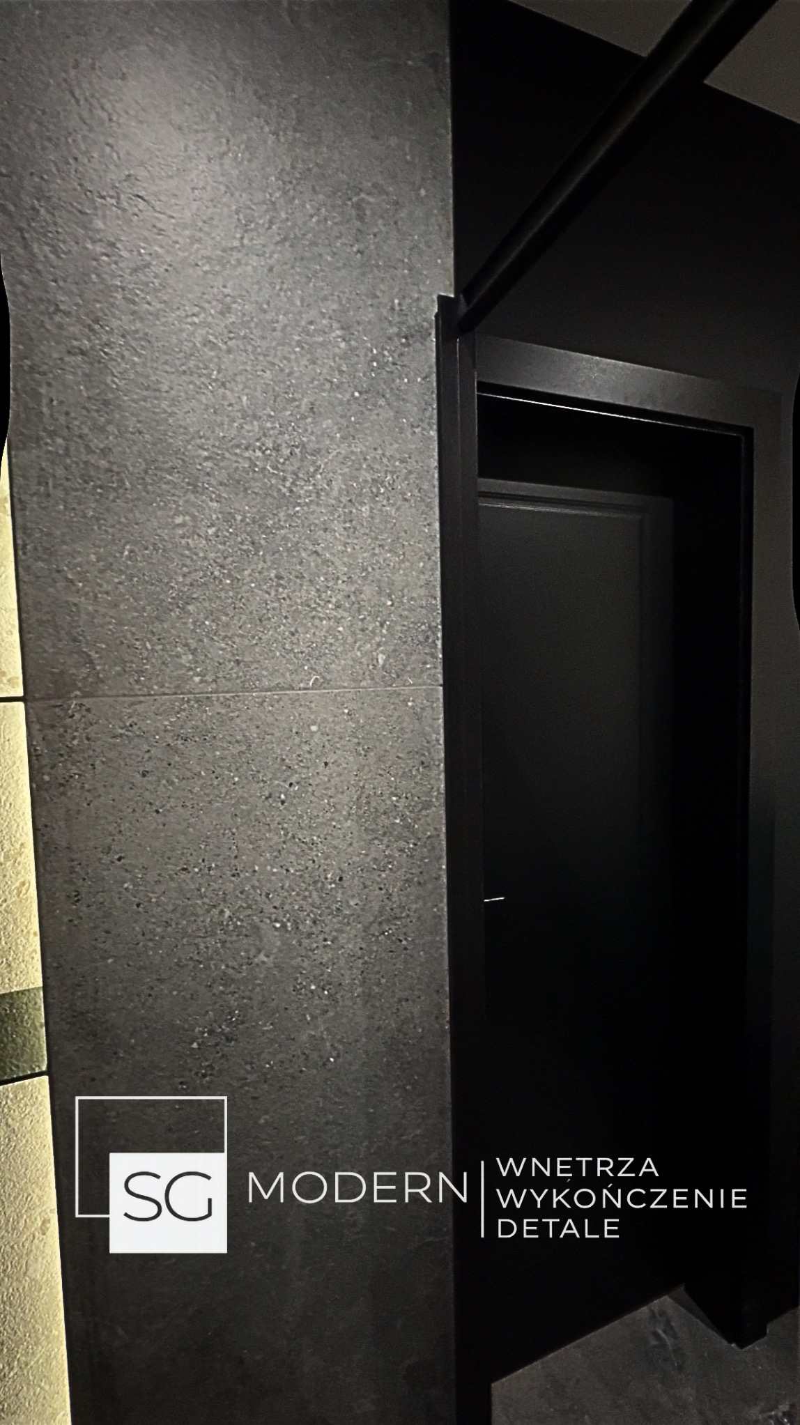 Minimalistyczne wnętrze: ciemnoszara ściana z płytek imitujących kamień, czarne drzwi wewnętrzne z ościeżnicą. Nowoczesny design z dbałością o detale.