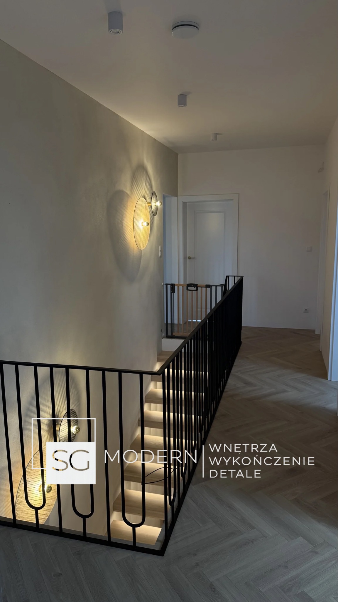 Nowoczesne wnętrze: schody z podświetleniem LED, czarna balustrada, parkiet w jodełkę. Na ścianie okrągły kinkiet z ażurowym kloszem. Minimalistyczny design.