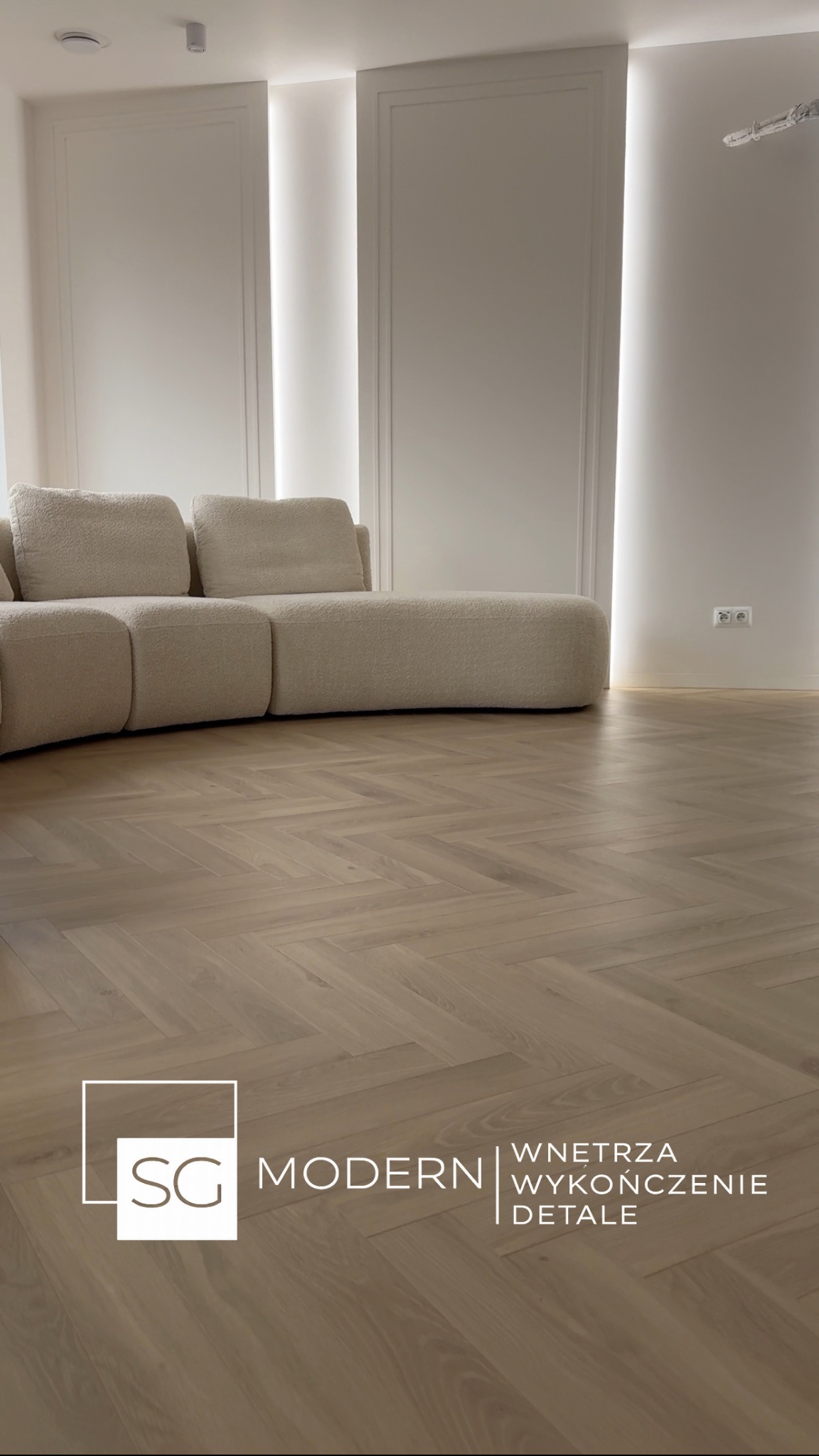 Jasny parkiet ułożony we wzór jodełki francuskiej, z widoczną sofą w tle i oświetleniem LED w ścianie. Minimalistyczne wnętrze z dbałością o detale.