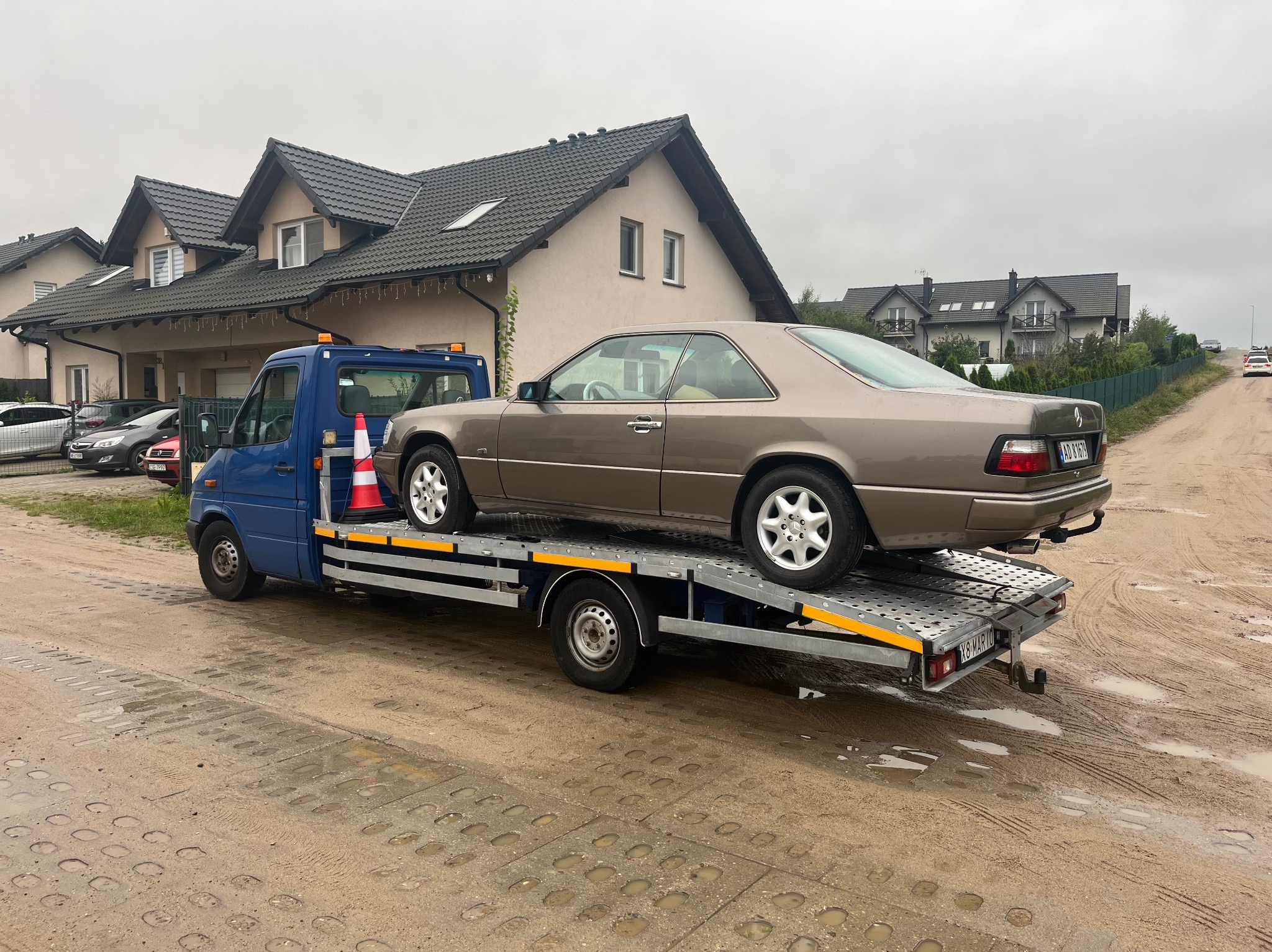 Brązowe auto Mercedes-Benz Coupe na niebieskiej platformie transportowej z pomarańczowym pachołkiem, na tle domów jednorodzinnych i szarego nieba.