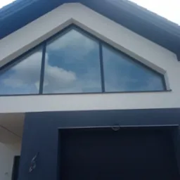 Nowoczesna elewacja domu jednorodzinnego z dużym tr&oacute;jkątnym oknem, odbijającym błękitne niebo z chmurami, kontrastująca z ciemnoszarą częścią garażową i jasną g&oacute;rną partią ściany.