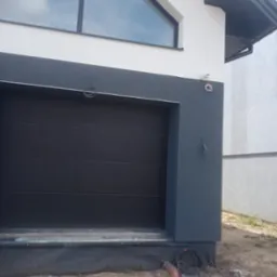 Dwukolorowa elewacja domu z czarną bramą garażową i oknem w kształcie trójkąta, widoczna kamera monitoringu i wystające kable przy fundamencie.