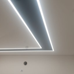 Tom-Kaj - Biały, błyszczący sufit z wbudowanym, geometrycznym oświetleniem LED w kształcie litery L i elementami drewnianymi, widok z dołu.