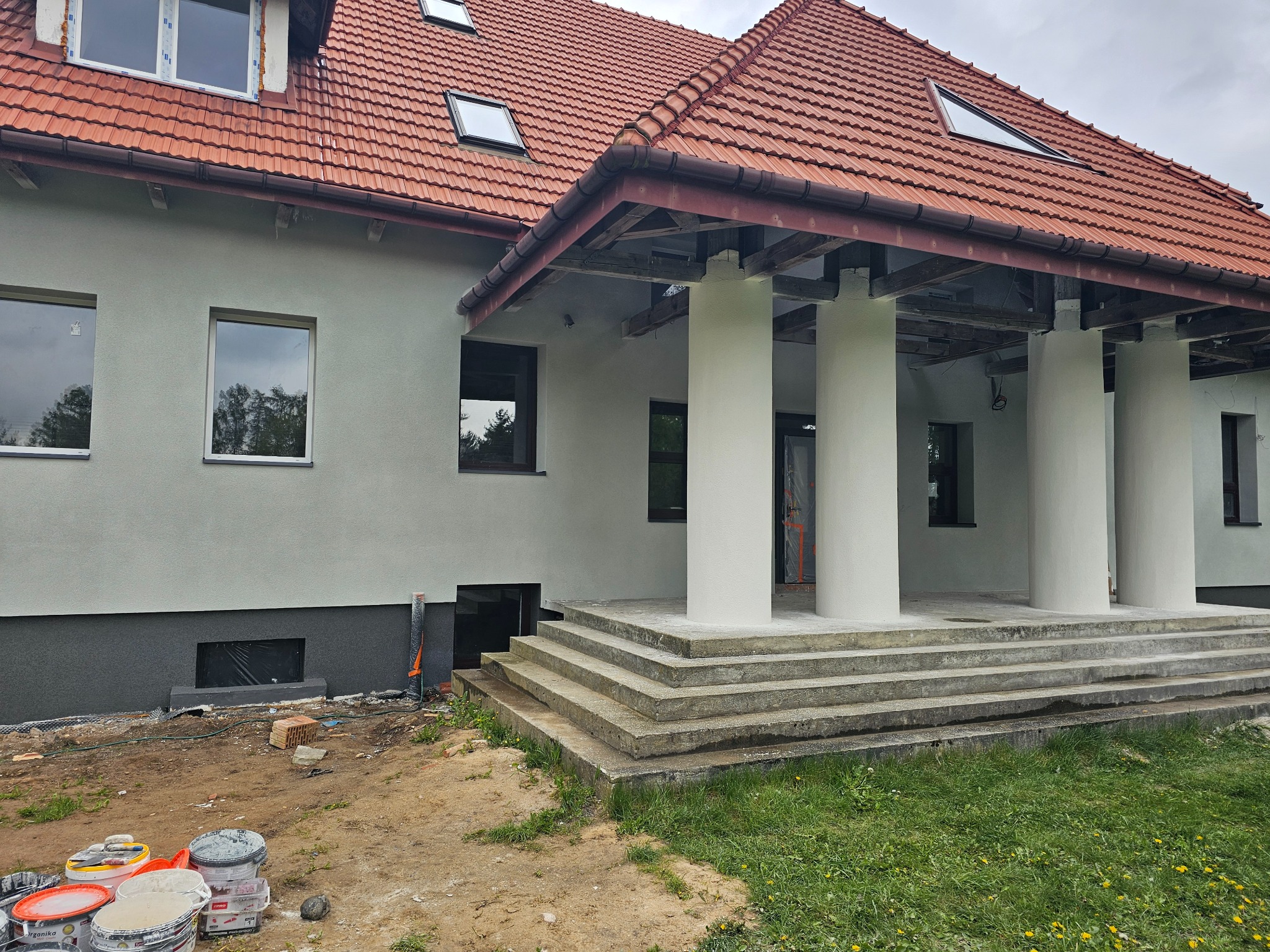 Dom jednorodzinny z nową, jasnoszarą elewacją i czerwonym dachem, widoczne kolumny przy wejściu, teren wokół domu w trakcie prac wykończeniowych, widoczne wiadra z farbą.