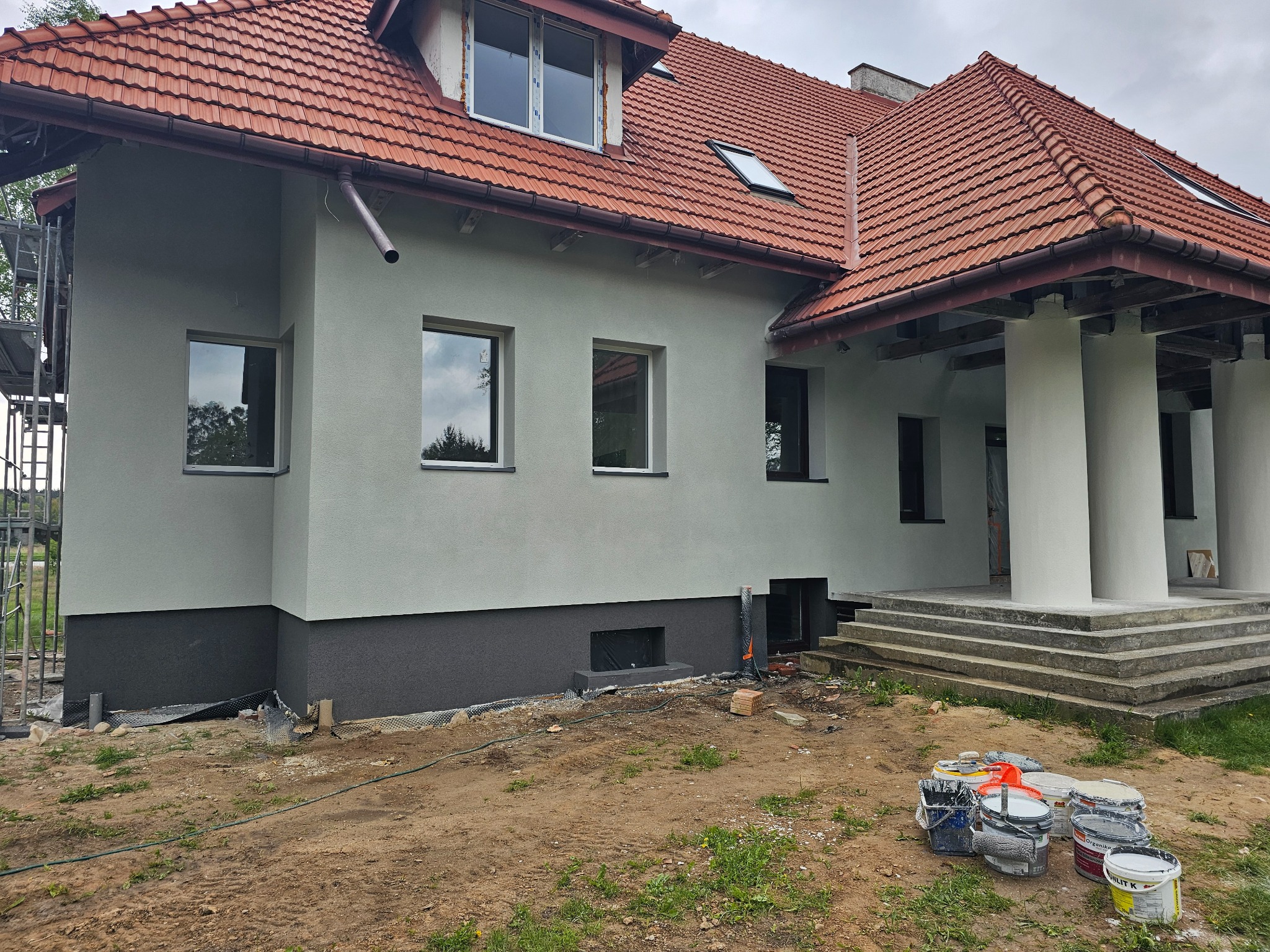 Dom jednorodzinny z nową, jasnoszarą elewacją i ciemnoszarym cokołem, widoczne puszki z farbą i rusztowanie w tle, prace w toku.