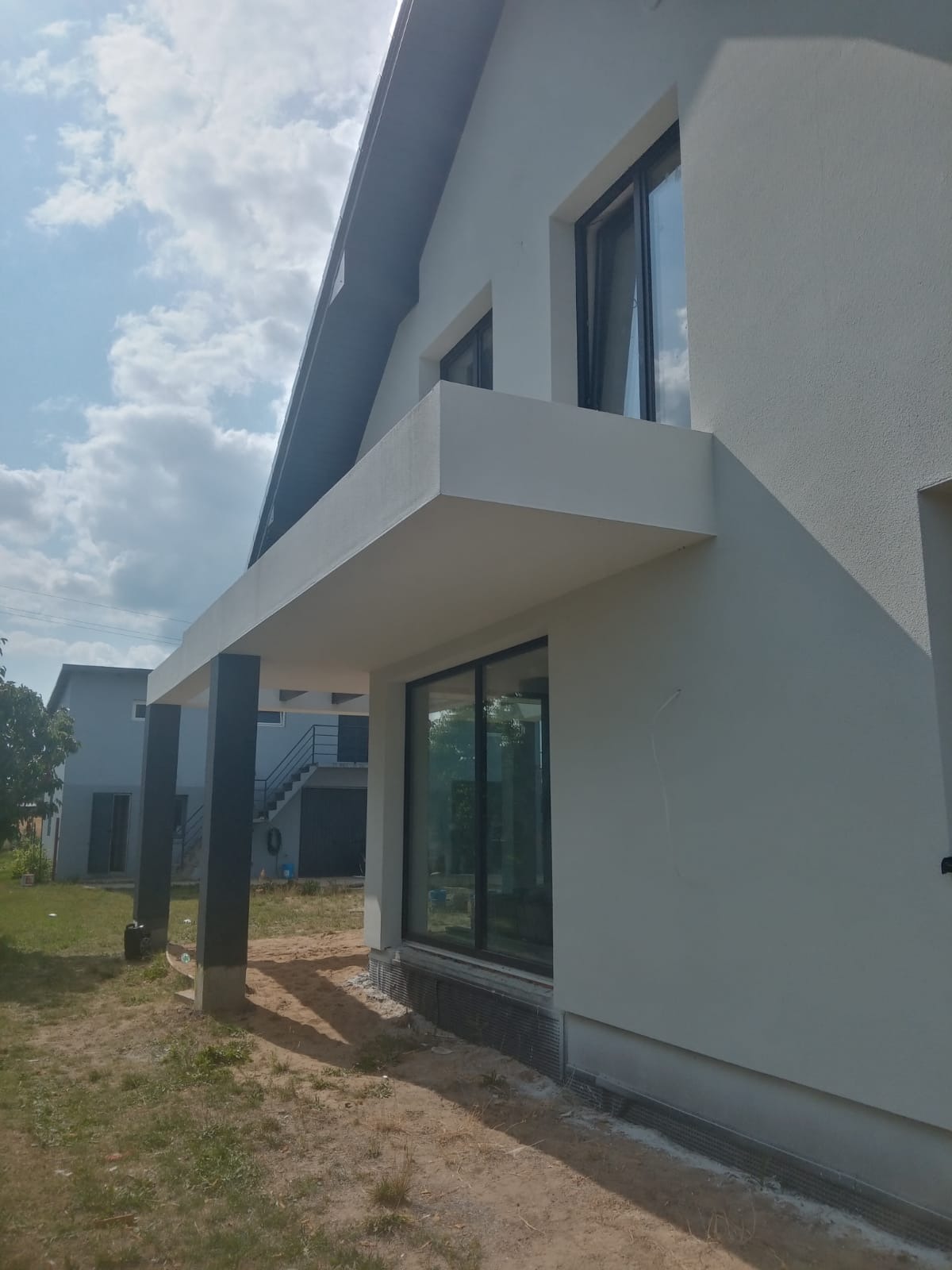 Świeżo otynkowana elewacja domu jednorodzinnego w Głownie, z widocznymi dużymi oknami i minimalistyczną bryłą budynku, w tle drugi budynek z zewnętrznymi schodami.