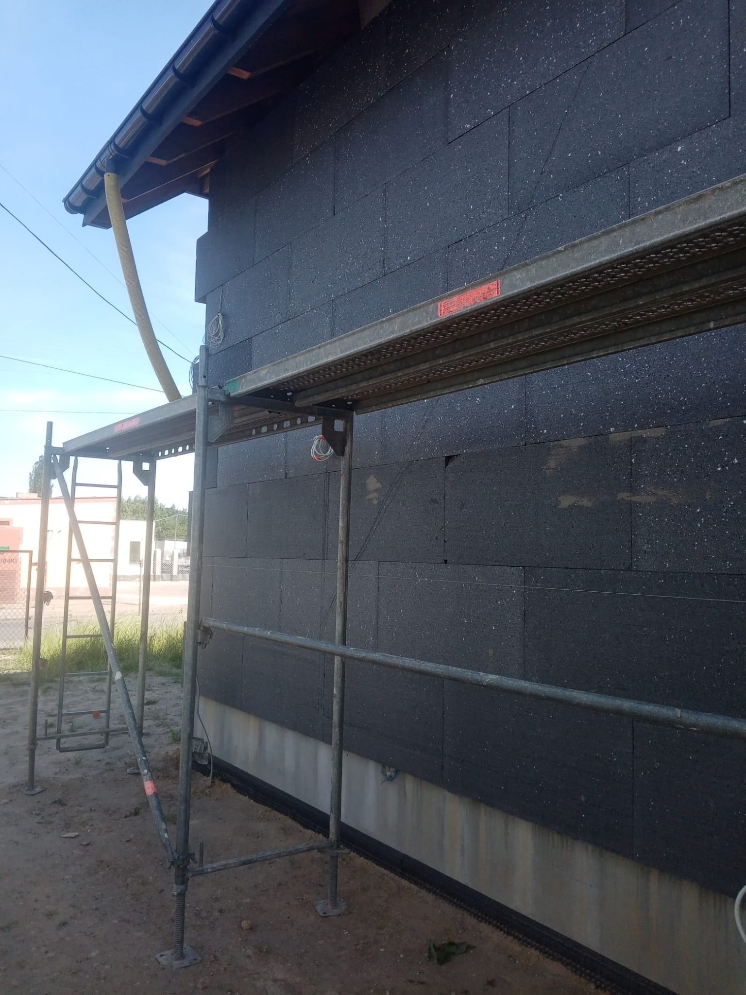 Ściana budynku pokryta czarnymi płytami izolacyjnymi, widoczne rusztowanie i rura wentylacyjna, przygotowania do dalszych prac wykończeniowych.