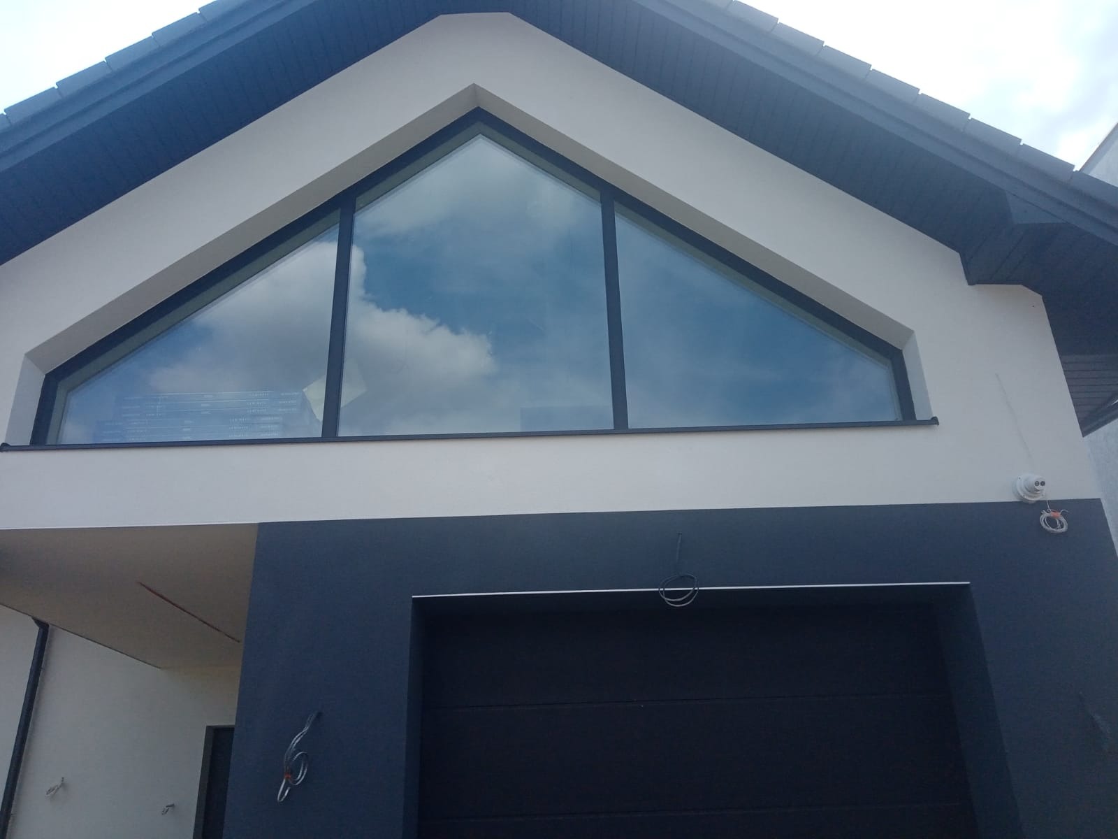 Nowoczesna elewacja domu jednorodzinnego z dużym trójkątnym oknem, odbijającym błękitne niebo z chmurami, kontrastująca z ciemnoszarą częścią garażową i jasną górną partią ściany.