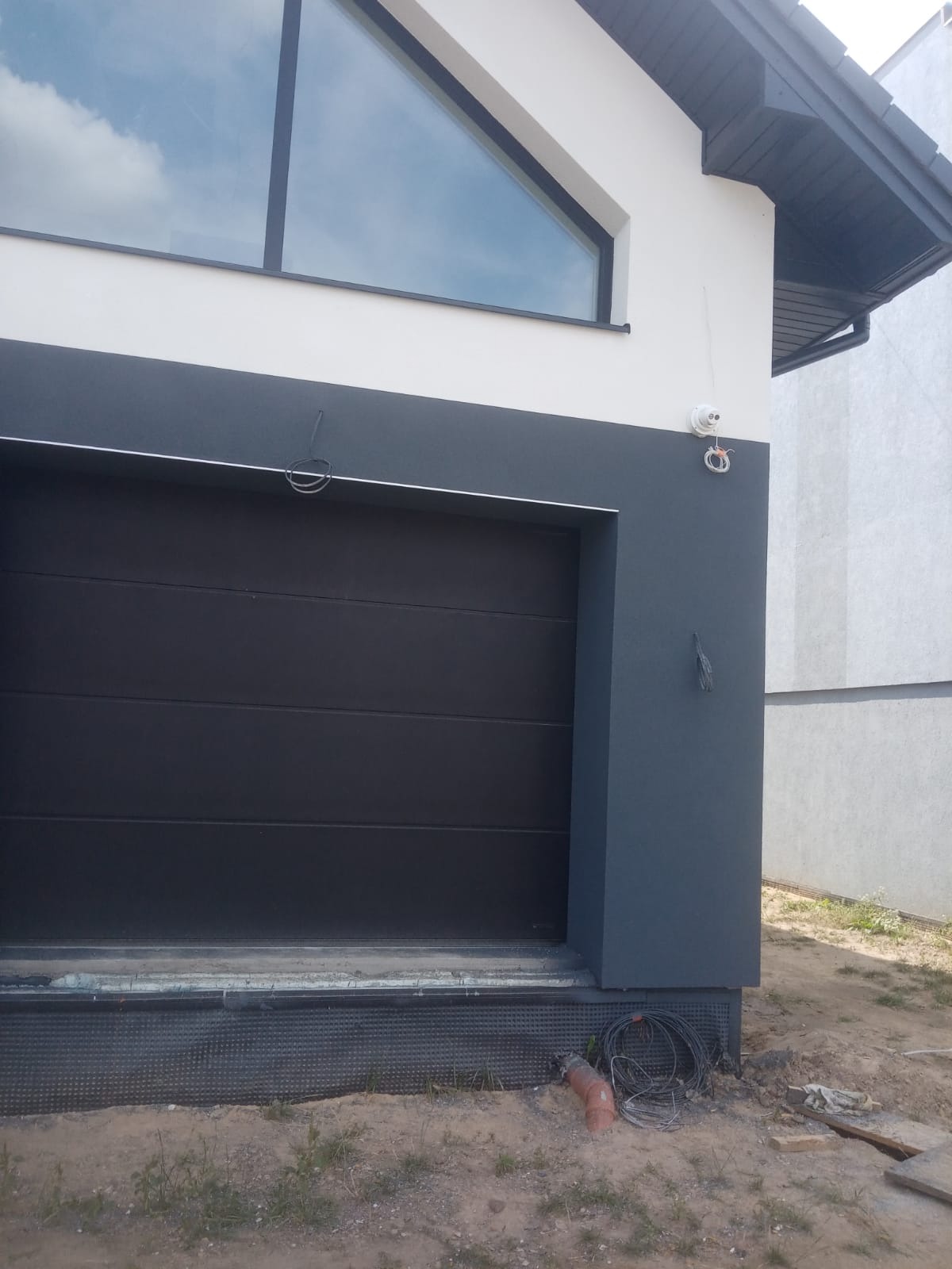 Dwukolorowa elewacja domu z czarną bramą garażową i oknem w kształcie trójkąta, widoczna kamera monitoringu i wystające kable przy fundamencie.