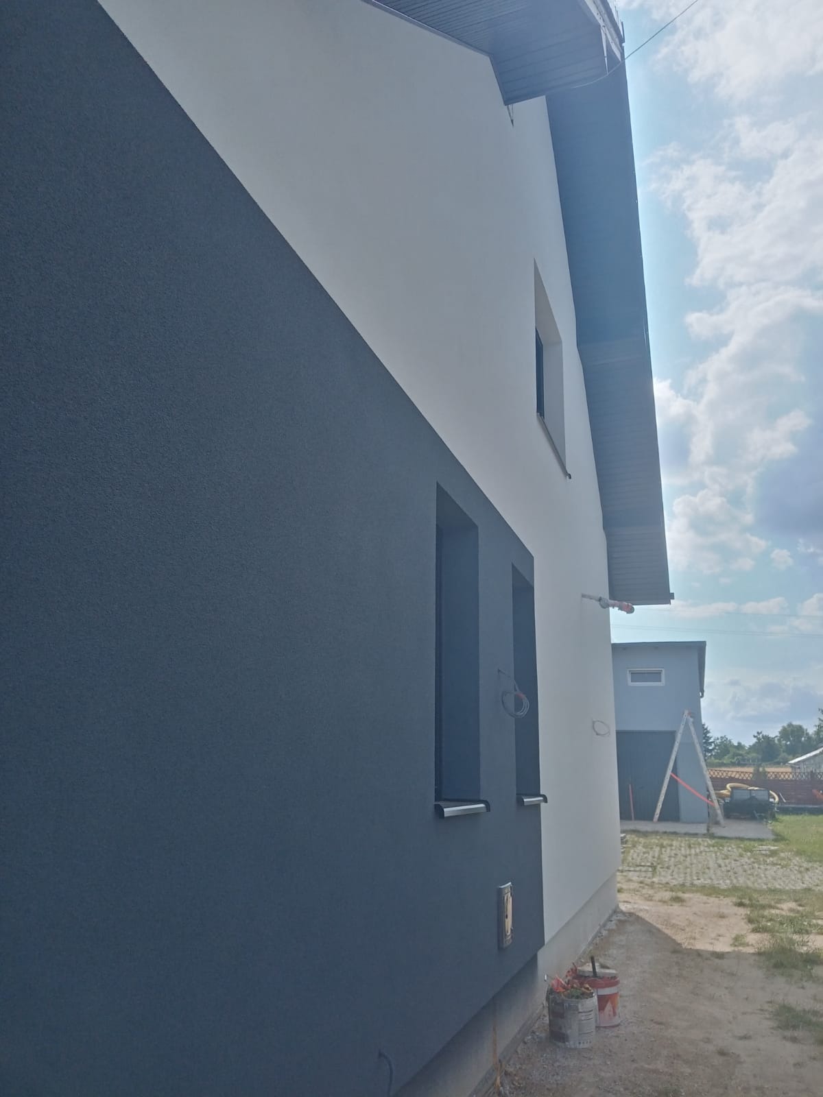 Dwukolorowa elewacja domu jednorodzinnego, z szarą częścią pokrytą wyraźną fakturą tynku i białym fragmentem, widoczne okna w szarych obramowaniach, w tle drugi budynek i drabina oparta o jego ścianę.