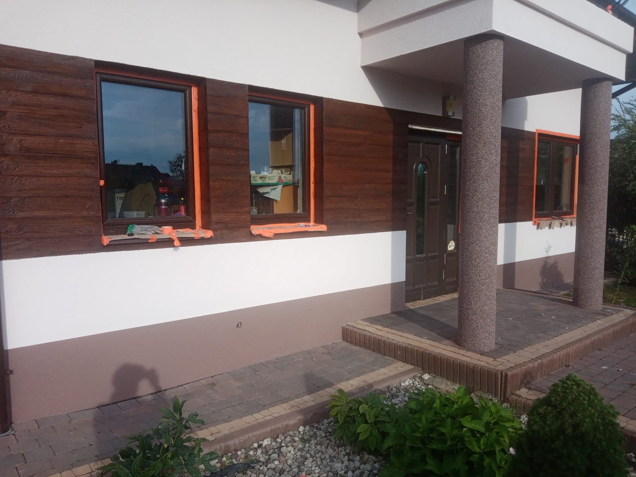 Elewacja domu z drewnopodobną okładziną, okna zabezpieczone taśmą malarską, widoczne narzędzia i pudełka wewnątrz, świeżo pomalowany cokół w kolorze brązowym.