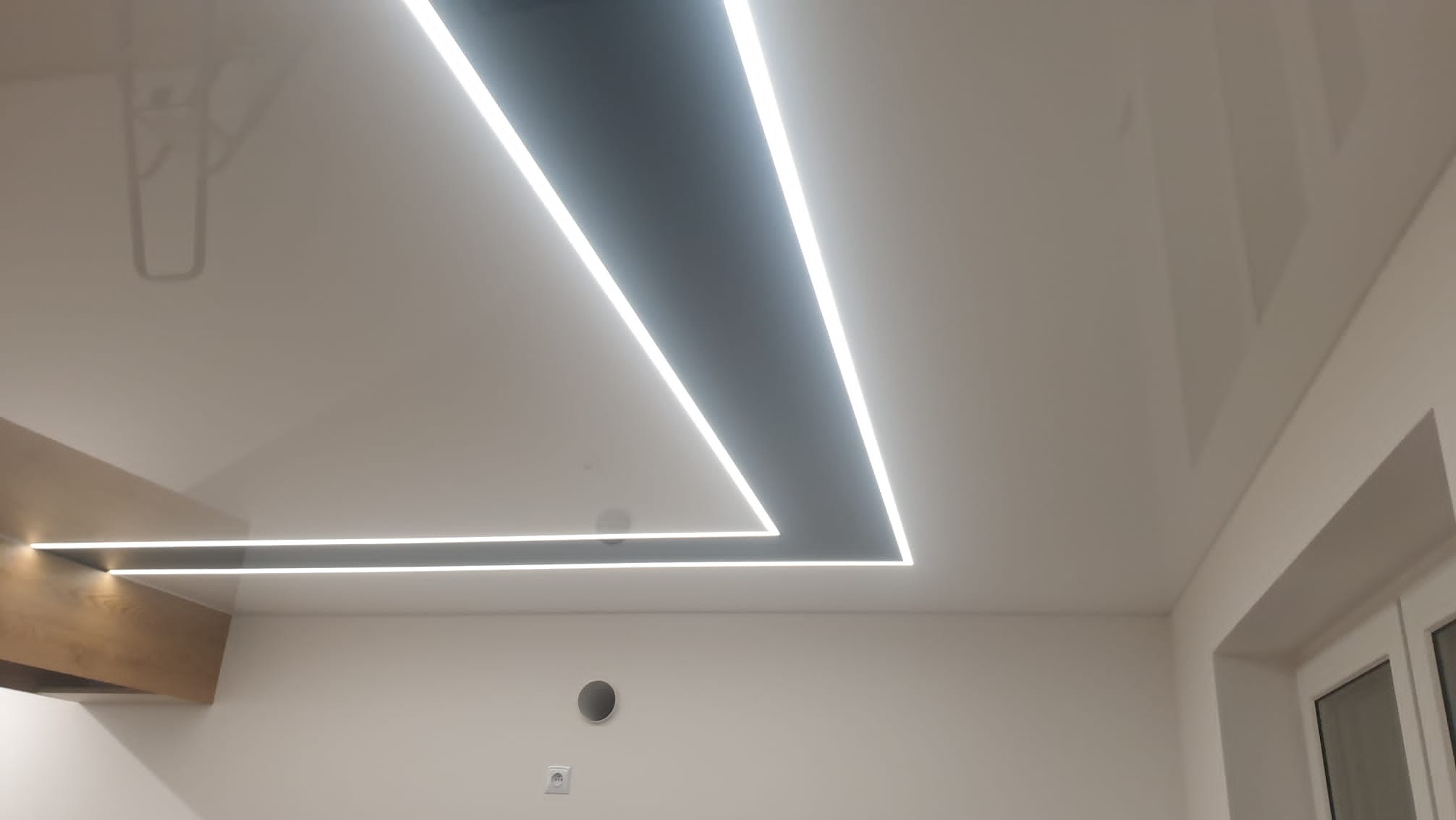Biały, błyszczący sufit z wbudowanym, geometrycznym oświetleniem LED w kształcie litery L i elementami drewnianymi, widok z dołu.