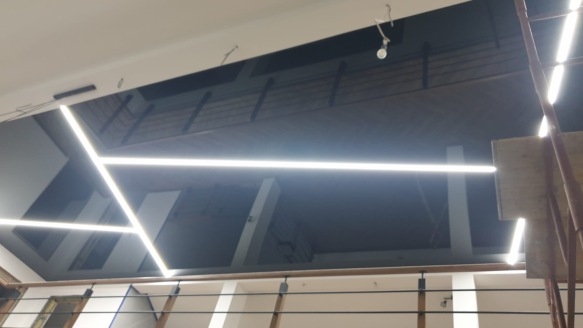 Sufit podwieszany z czarną przestrzenią i geometrycznym oświetleniem LED, widok z dołu, fragment rusztowania po prawej stronie.