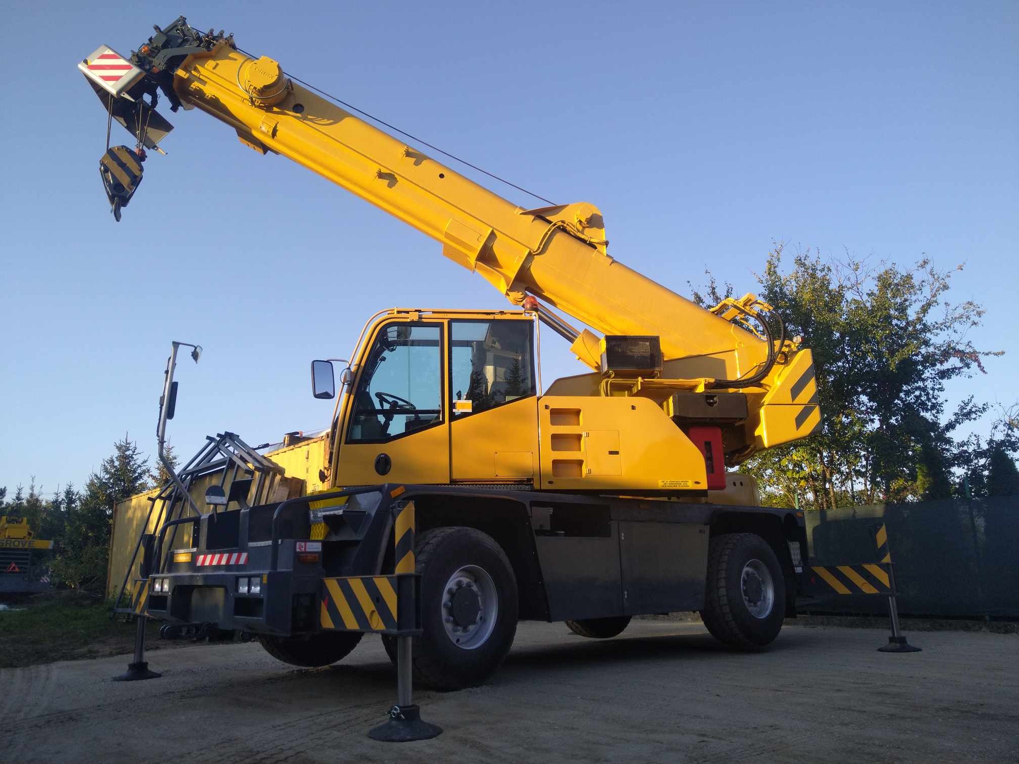 Żółty samojezdny żuraw teleskopowy Grove na rozłożonych podporach, z wysuniętym ramieniem i hakiem, na tle niebieskiego nieba i zieleni.