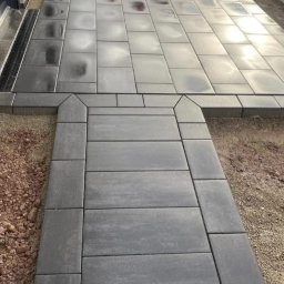 Stachu - Ułożona, szara kostka brukowa prowadząca do domu. Widoczny fragment elewacji, zaparkowane samochody i koparka Kubota w tle. Kostka ułożona w regularny wzór.