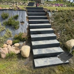 Stachu - Schody z płyt betonowych prowadzące w górę zbocza, z widoczną łopatą i taczką na szczycie. Elementy krajobrazu: kamienie, roślinność, folia ochronna.