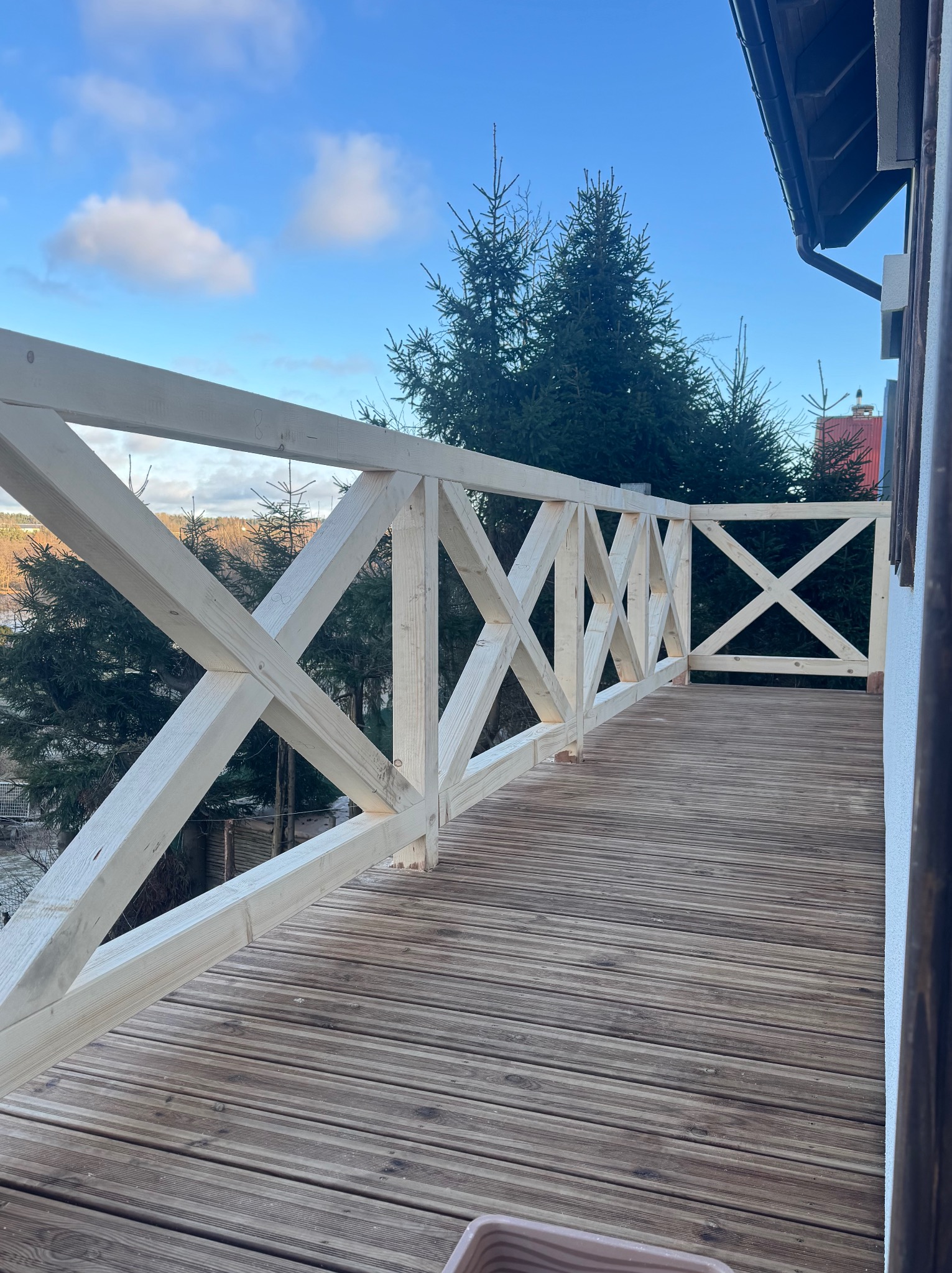 Jasny, drewniany taras z balustradą w kształcie krzyży, otoczony zielenią drzew. Widok na błękitne niebo z chmurami. Elementy konstrukcyjne w surowym stanie.