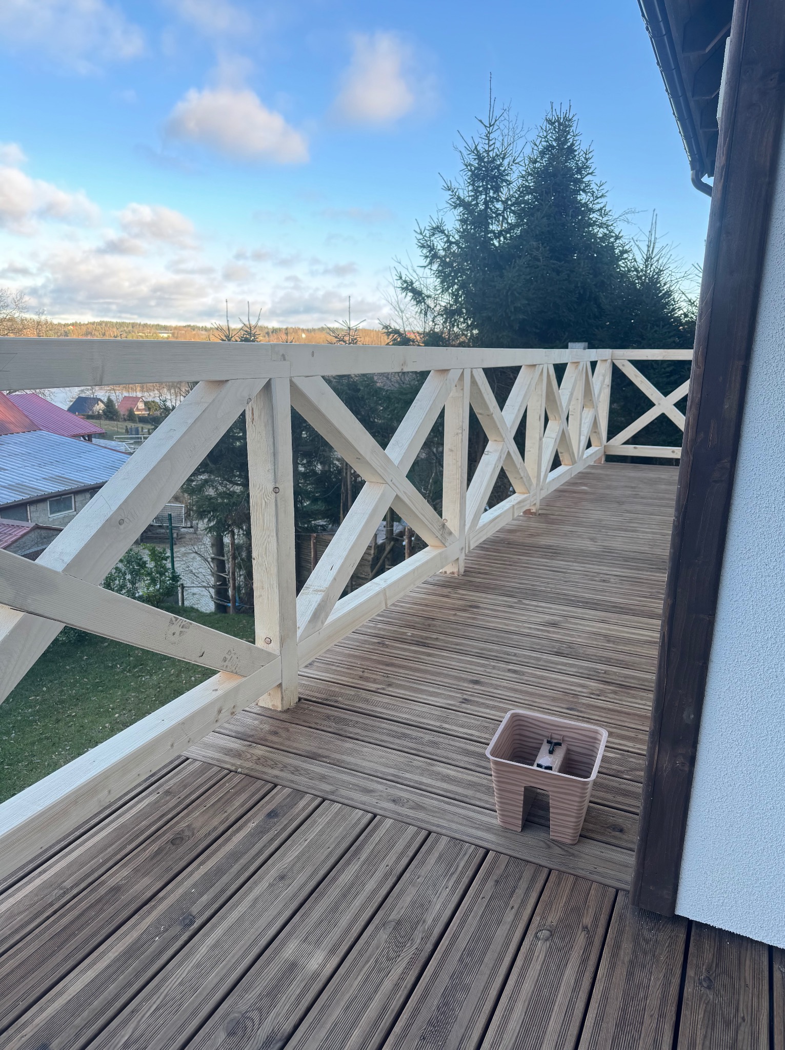 Nowy taras drewniany z białymi, krzyżowymi barierkami i widokiem na jezioro. Doniczka na podłodze. Jasne, naturalne światło.