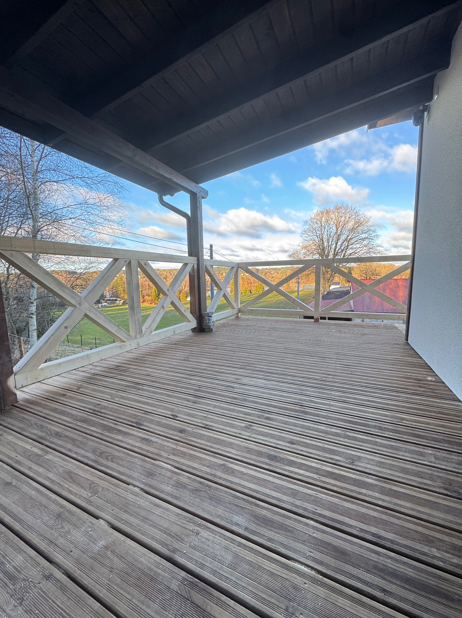 Zadaszony taras drewniany z jasną, drewnianą balustradą krzyżową. Widok na zewnątrz, błękitne niebo i drzewa. Deski tarasowe ułożone poziomo, widoczne słoje drewna.