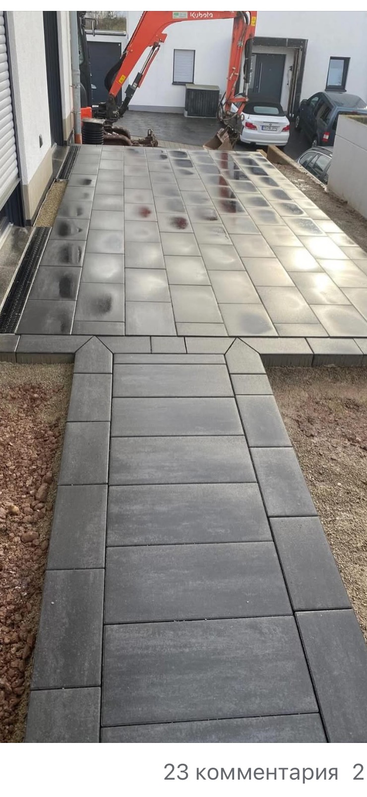 Ułożona, szara kostka brukowa prowadząca do domu. Widoczny fragment elewacji, zaparkowane samochody i koparka Kubota w tle. Kostka ułożona w regularny wzór.