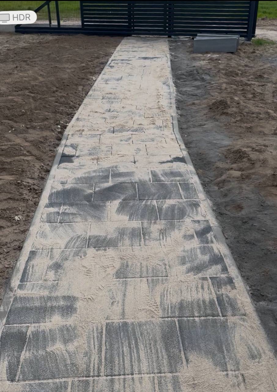 Ścieżka z szarej kostki brukowej, świeżo po zasypaniu piaskiem, prowadząca do czarnego ogrodzenia panelowego. Widoczne ślady stóp na piasku.