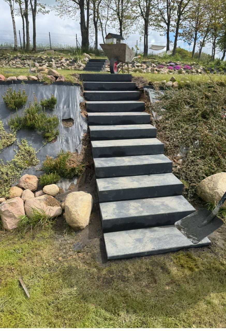 Schody z płyt betonowych prowadzące w górę zbocza, z widoczną łopatą i taczką na szczycie. Elementy krajobrazu: kamienie, roślinność, folia ochronna.