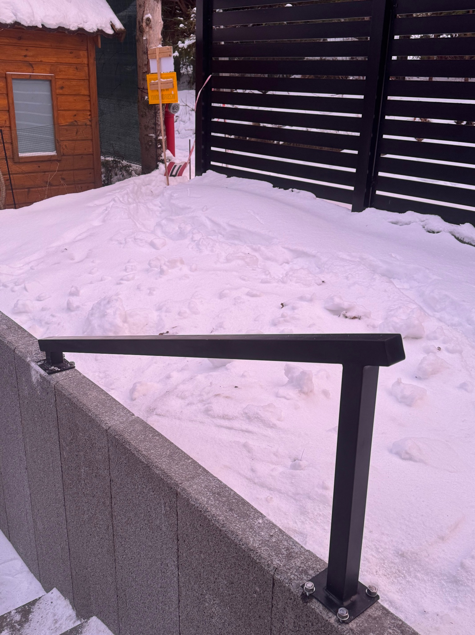 Nowoczesne, poziome ogrodzenie panelowe w kolorze czarnym, zamontowane na betonowym murku w zimowej scenerii. Widoczne śruby mocujące.