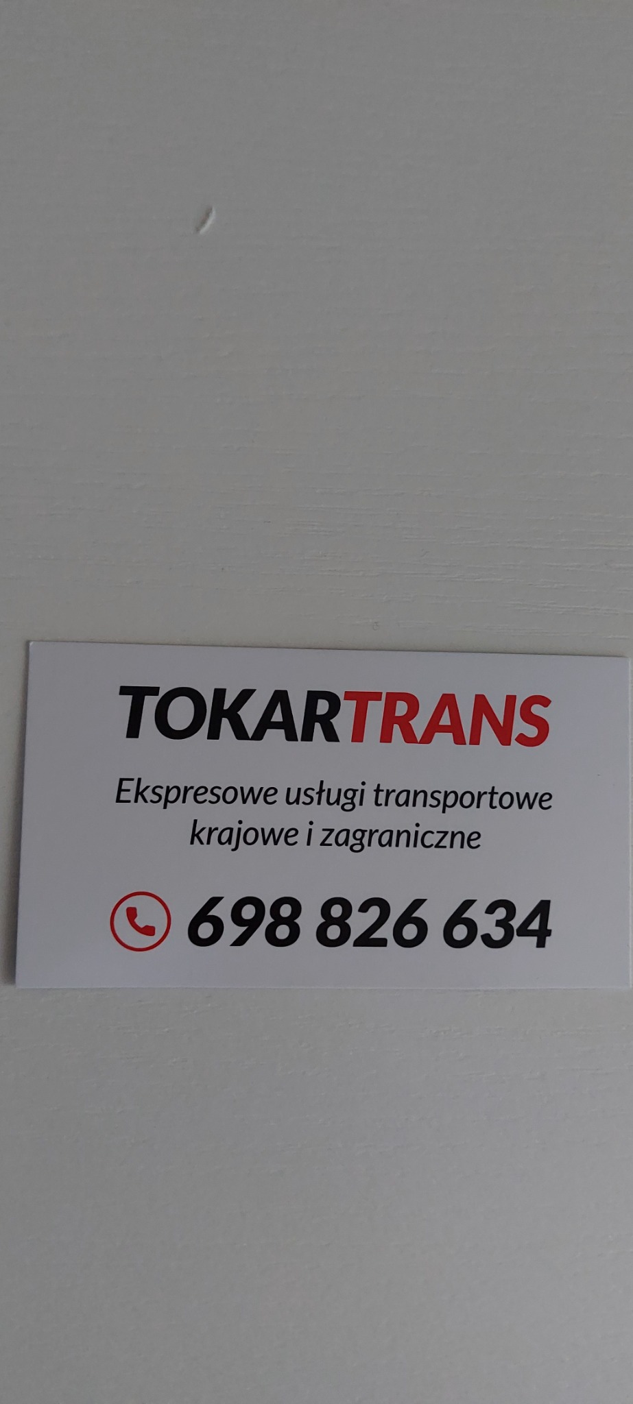 Wizytówka firmy TokarTrans z numerem telefonu i informacją o ekspresowych usługach transportowych krajowych i zagranicznych na białym tle ściany.