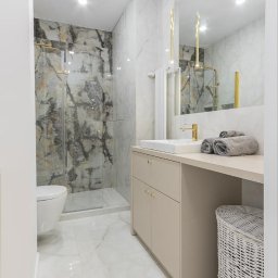 Oleksandr Nazarow - Jasna łazienka z marmurowymi płytkami w odcieniach szarości i złota, kabina prysznicowa ze złotymi okuciami, beżowa szafka z umywalką i złotą armaturą, dekoracyjne lampy wiszące, biała podłoga...