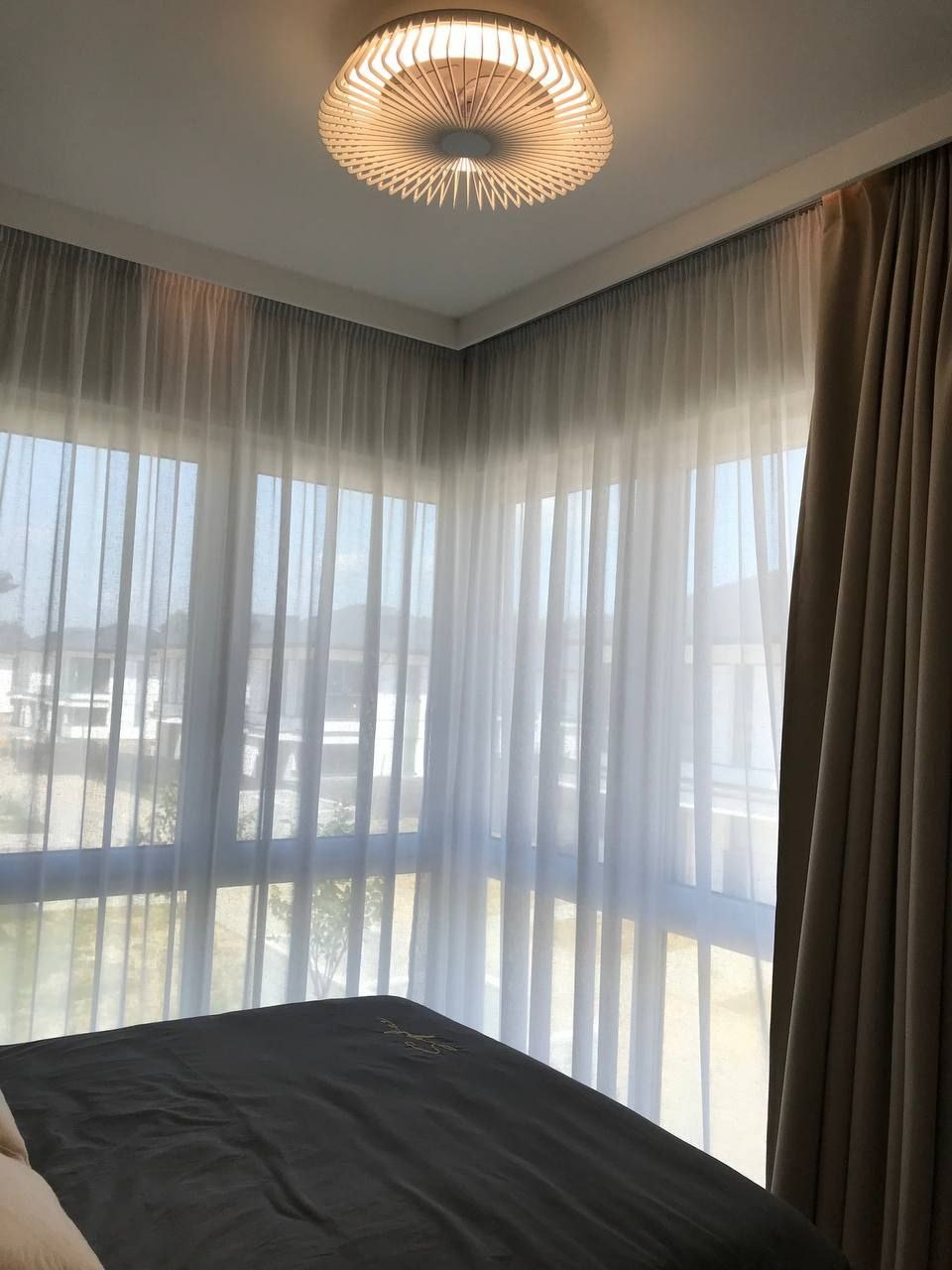 Sypialnia z łóżkiem w szarym odcieniu, zasłonięta oknami z delikatnymi, białymi firanami i ciężkimi, beżowymi zasłonami. Na suficie okrągła, nowoczesna lampa z drewnianymi elementami. Widok z okna...