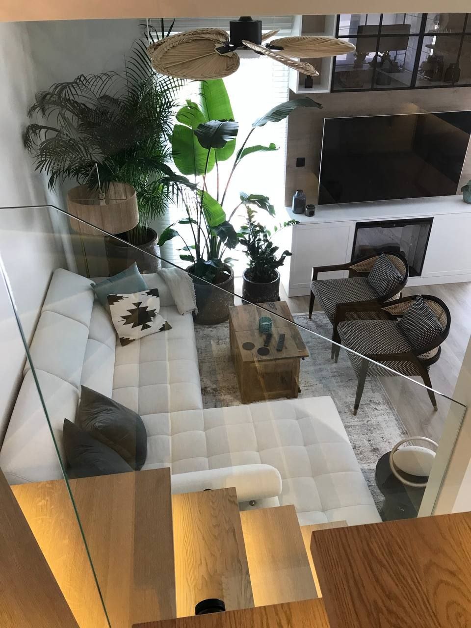 Jasny salon z białym narożnikiem, drewnianym stolikiem kawowym i licznymi roślinami doniczkowymi, widok z góry przez szklaną balustradę schodów.