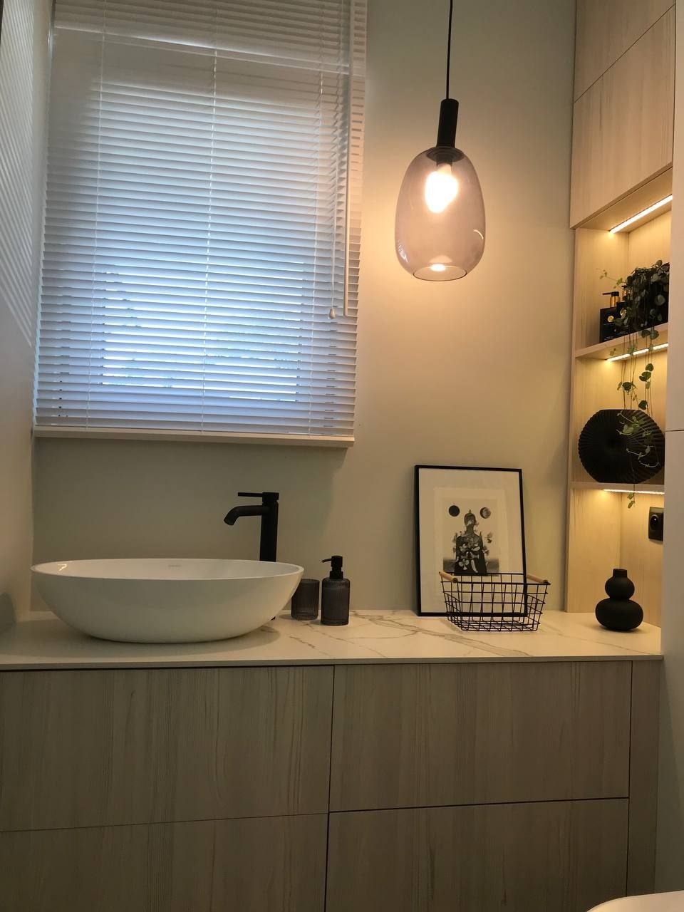 Nowoczesna łazienka z owalną umywalką nablatową, czarną armaturą, blatem imitującym marmur, szafkami z jasnego drewna oraz wiszącą lampą z przydymionego szkła. Na półkach dekoracyjne rośliny...