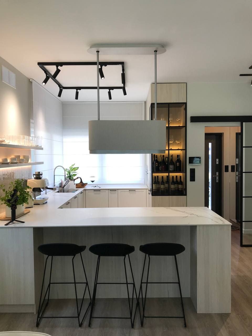 Nowoczesna kuchnia z wyspą, trzema czarnymi stołkami barowymi, oświetleniem punktowym na suficie i przeszkloną szafką z winami w tle.