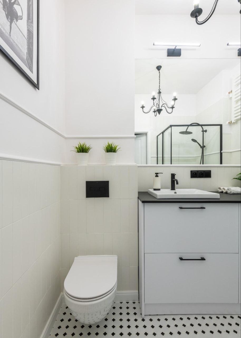 Biała łazienka z czarnymi akcentami: umywalka z szafką, toaleta, płytki podłogowe w czarno-białą szachownicę, lustro odbijające kabinę prysznicową i czarny żyrandol.