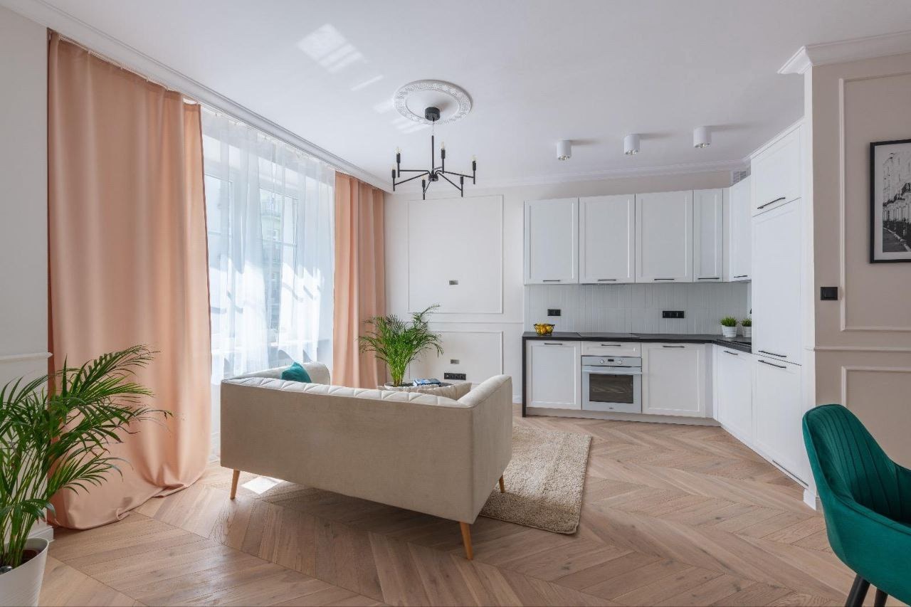 Jasny salon z aneksem kuchennym, parkiet ułożony w jodełkę, beżowa sofa z turkusową poduszką, białe szafki kuchenne z czarnym blatem, dekoracyjny żyrandol i sztukateria na suficie.