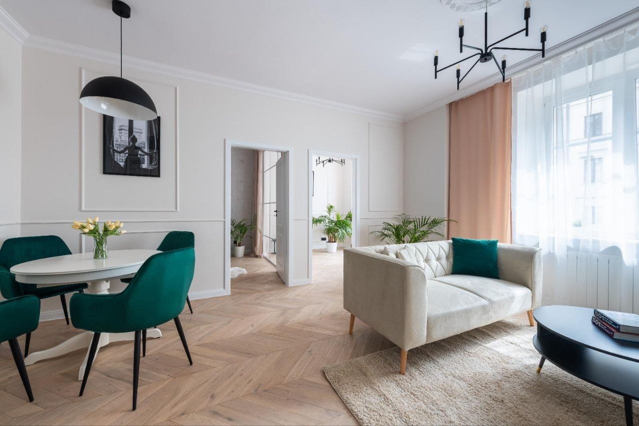 Jasne wnętrze z parkietem w jodełkę, okrągłym stołem z zielonymi krzesłami, kremową sofą z zieloną poduszką i czarnym stolikiem kawowym, doświetlone naturalnym światłem z okna.