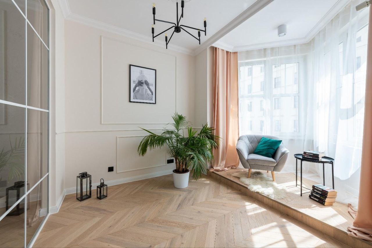 Jasny salon z parkietem w jodełkę, kremowymi ścianami z dekoracyjnymi panelami, nowoczesnym czarnym żyrandolem, fotelem z zieloną poduszką w wykuszowej wnęce okiennej z jasnymi zasłonami i rośliną...