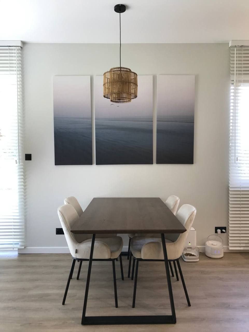 Jasna jadalnia z drewnianym stołem na metalowych nogach, welurowe krzesła, bambusowa lampa wisząca i tryptyk z motywem morza na ścianie, widok z przodu, automatyczny podajnik karmy dla zwierząt...