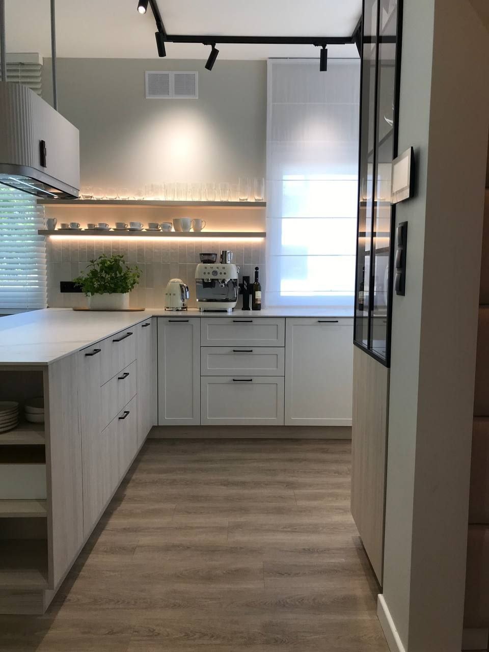 Nowoczesna kuchnia z białymi szafkami z ciemnymi uchwytami, drewnianą podłogą, oświetleniem LED pod półką z filiżankami i szklankami, oraz ekspresem do kawy.