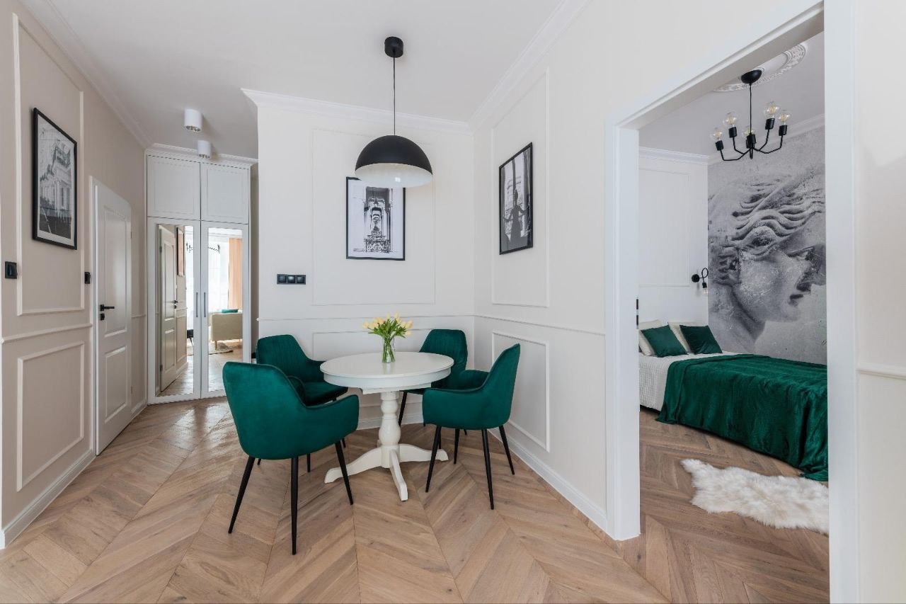 Jasne wnętrze z parkietem w jodełkę, okrągłym stołem z bukietem tulipanów i czterema szmaragdowymi krzesłami, widok na sypialnię z fototapetą przedstawiającą grecką rzeźbę.