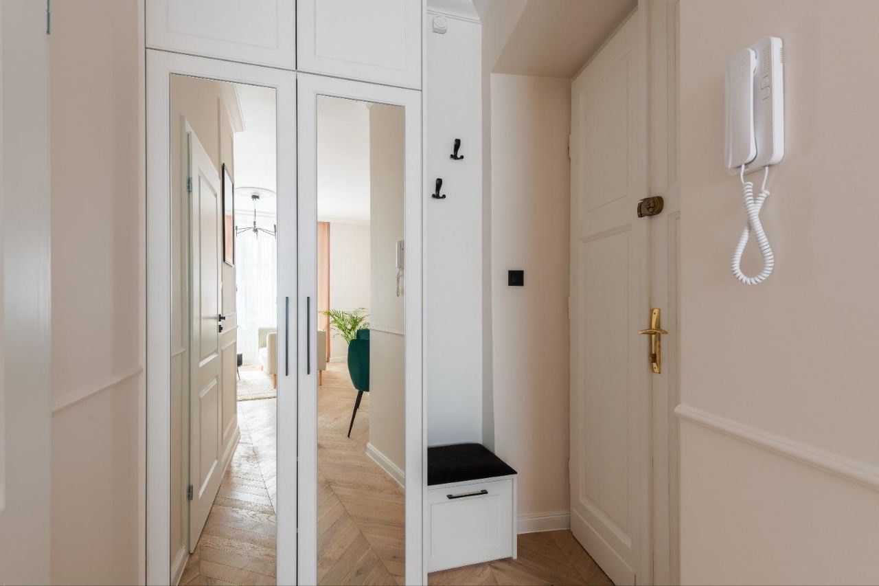 Biały przedpokój z lustrzaną szafą wnękową, parkietem w jodełkę i siedziskiem z czarną poduszką, widok na dalszą część mieszkania przez otwarte drzwi.