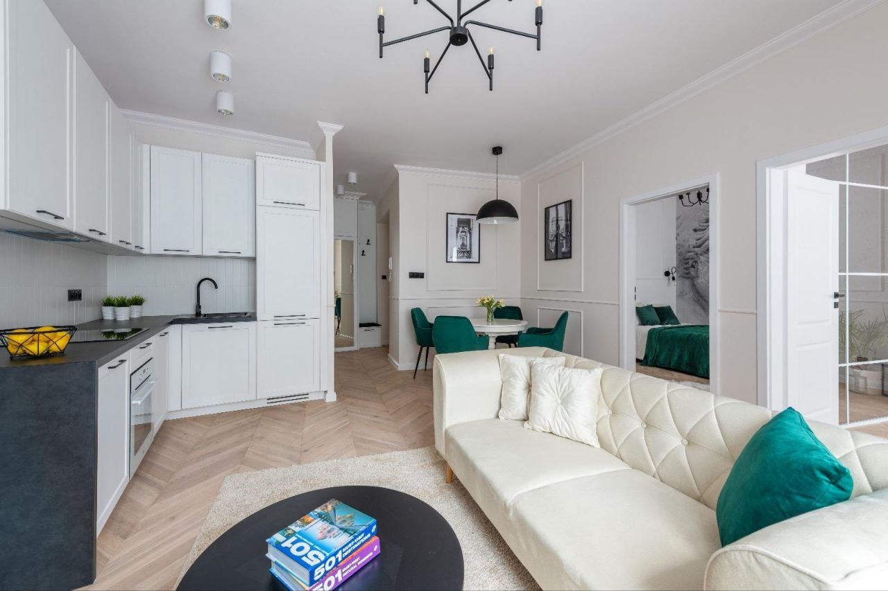 Jasne, przestronne wnętrze po remoncie z białymi szafkami kuchennymi, parkietem w jodełkę, kremową sofą z zielonymi akcentami i okrągłym stolikiem kawowym z książkami, widok na jadalnię i sypialnię...