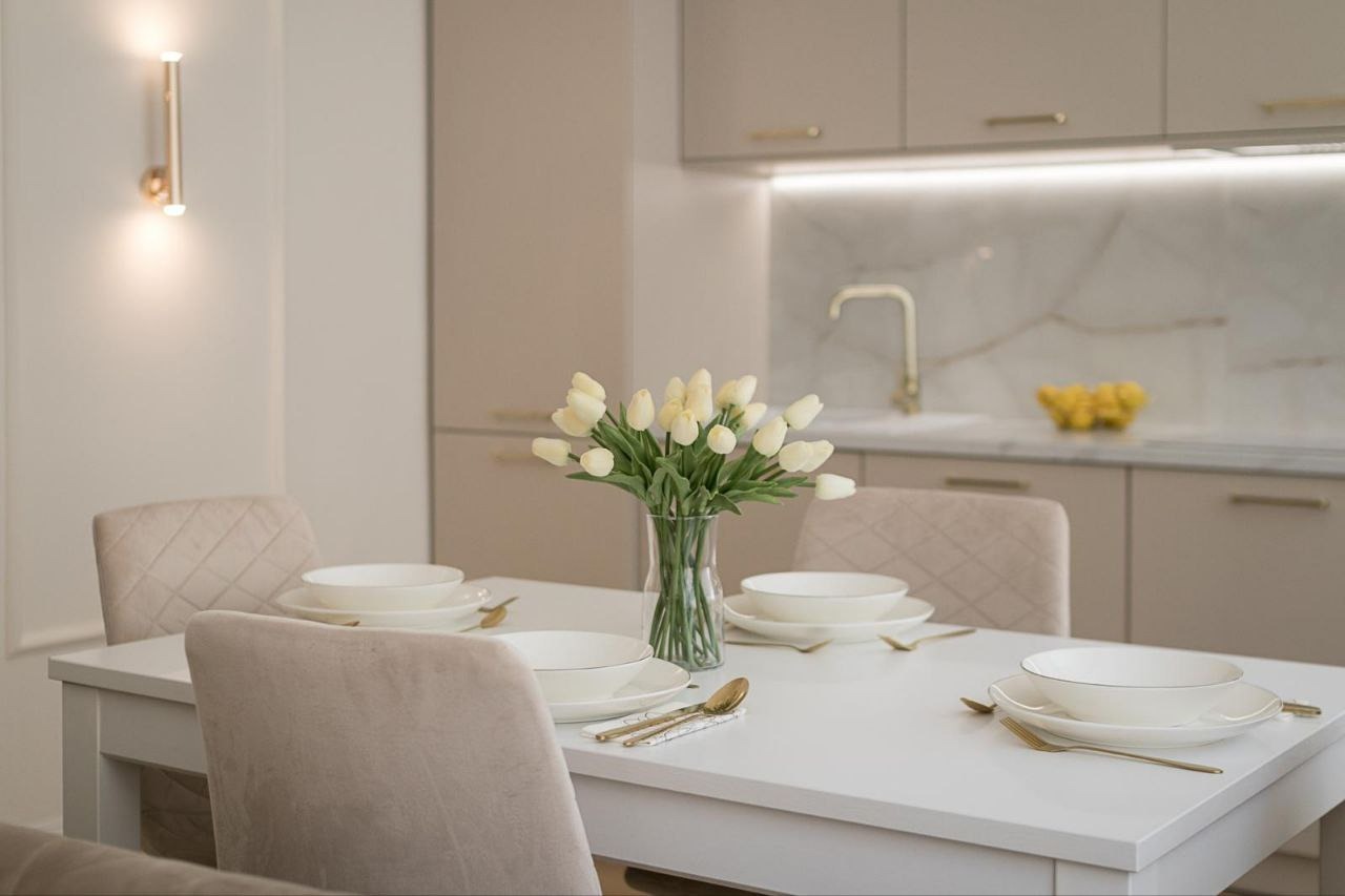 Elegancka, beżowa kuchnia z marmurowym blatem i złotymi akcentami, stół z nakryciem i wazonem tulipanów w centrum kadru.