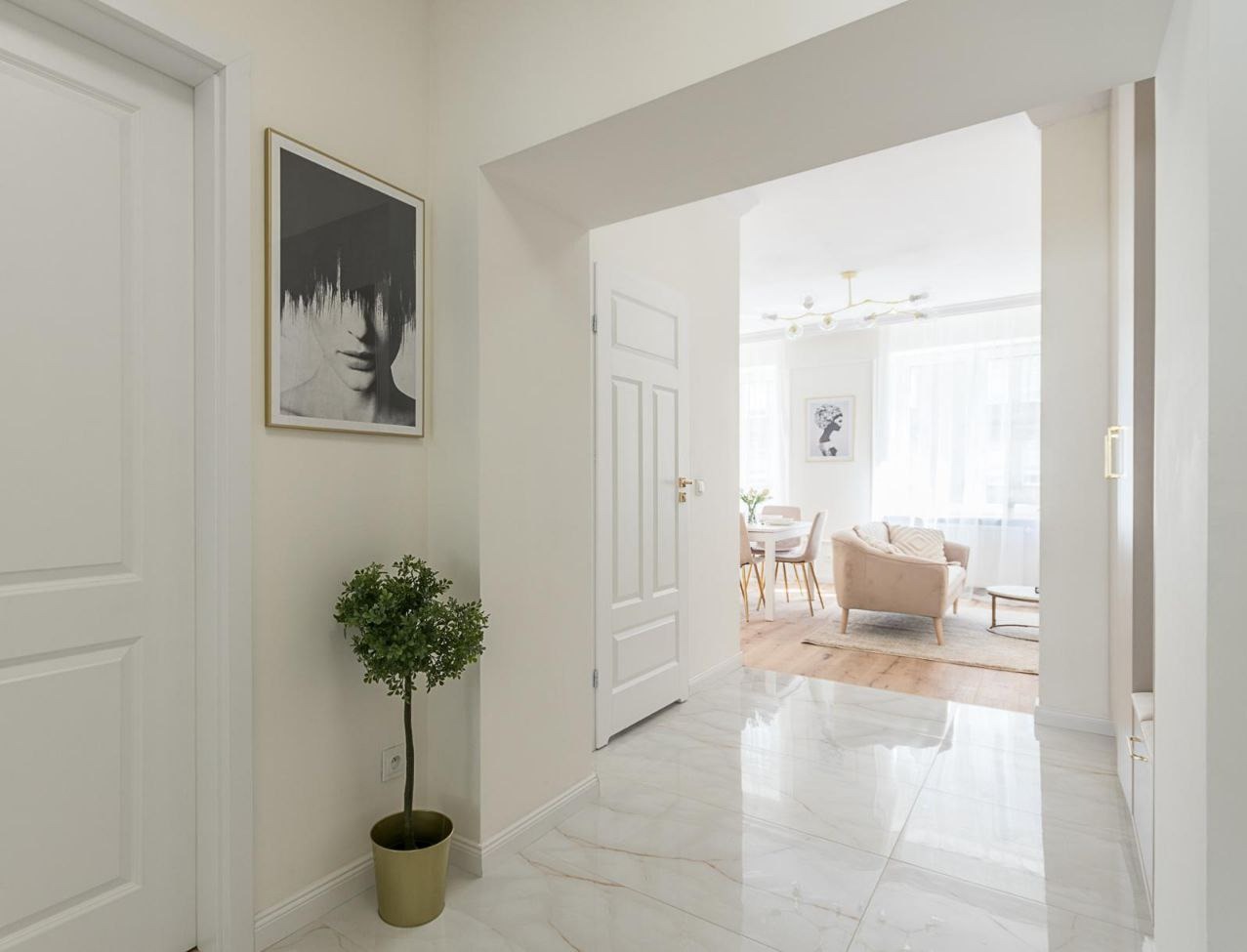 Jasne wnętrze mieszkania z białymi drzwiami, marmurową podłogą, sztuką na ścianie i widokiem na salon z beżową sofą
